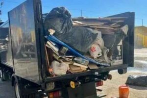 A black garbage truck with its back open, revealing an overfilled load of trash bags, wooden planks, and assorted waste. Some debris is spilling out the back, and there is a red bucket on the ground nearby.